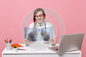 Female doctor sit at desk work on computer with medical document hold X-ray in hospital isolated on pastel pink wall