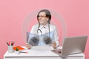 Female doctor sit at desk work on computer with medical document hold X-ray in hospital isolated on pastel pink wall