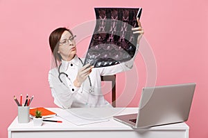 Female doctor sit at desk work on computer with medical document hold x-ray in hospital isolated on pastel pink wall