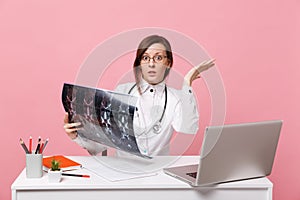 Female doctor sit at desk work on computer with medical document hold x-ray in hospital isolated on pastel pink wall