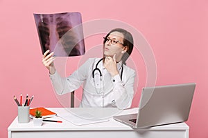 Female doctor sit at desk work on computer with medical document hold X-ray in hospital isolated on pastel pink wall