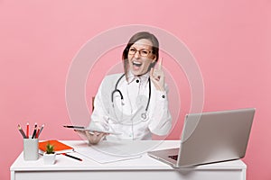 Female doctor sit at desk work on computer with medical document hold tablet in hospital isolated on pastel pink wall