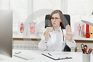Female doctor sitting at desk holding bundle of dollars cash money, say No to bribe, showing stop gesture with palm in
