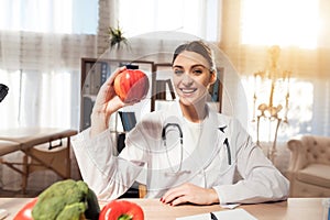 Female doctor sitting at desk in office with microscope and stethoscope. Woman is holding red apple.