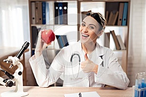 Female doctor sitting at desk in office with microscope and stethoscope. Woman is holding red apple.