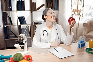Female doctor sitting at desk in office with microscope and stethoscope. Woman is holding red apple.