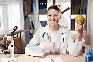 Female doctor sitting at desk in office with microscope and stethoscope. Woman is holding yellow apple.