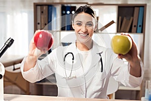 Female doctor sitting at desk in office with microscope and stethoscope. Woman is holding yellow and red apples.