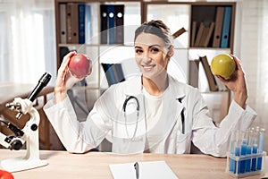 Female doctor sitting at desk in office with microscope and stethoscope. Woman is holding yellow and red apples.