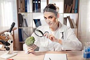 Female doctor sitting at desk in office with microscope and stethoscope. Woman is looking at broccoli with magnifyer.