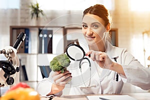 Female doctor sitting at desk in office with microscope and stethoscope. Woman is looking at broccoli with magnifyer.