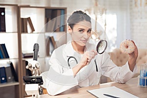 Female doctor sitting at desk in office with microscope and stethoscope. Woman is looking at donut with magnifyer.