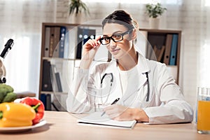 Female doctor sitting at desk in office with microscope and stethoscope. Woman is writing on clipboard.