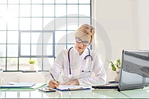 Female doctor sitting at desk and using laptop and doing some paperwork in doctorÃ¢â¬â¢s office photo