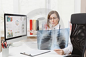 Female doctor sitting at desk, working on computer, holding X-ray of lungs, fluorography, roentgen in light office in