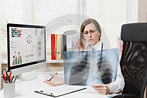 Female doctor sitting at desk, working on computer, holding X-ray of lungs, fluorography, roentgen in light office in