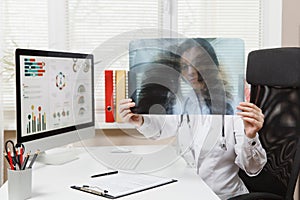 Female doctor sitting at desk, working on computer, holding X-ray of lungs, fluorography, roentgen in light office in