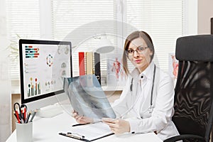 Female doctor sitting at desk, working on computer, holding X-ray of lungs, fluorography, roentgen in light office in