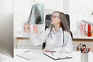 Female doctor sitting at desk, working on computer, holding X-ray of lungs, fluorography, roentgen in light office in