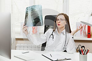 Female doctor sitting at desk, working on computer, holding X-ray of lungs, fluorography, roentgen in light office in