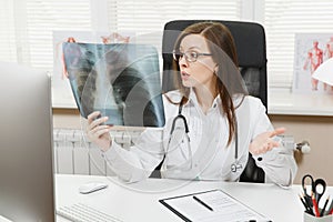 Female doctor sitting at desk, working on computer, holding X-ray of lungs, fluorography, roentgen in light office in