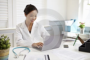 Female Doctor Sitting At Desk Working At Laptop In Office