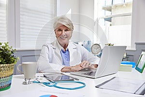 Female Doctor Sitting At Desk Working At Laptop In Office