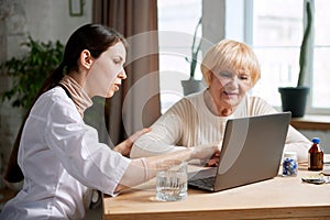 Female doctor sitting at table in office with her senior patient and talking, looking on laptop. Explaining medical