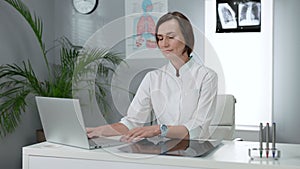 Female Doctor Sitting At The Table And Using Laptop. Young Woman In White Coat Sitting In The Office.