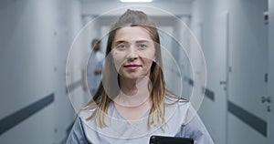 Female doctor stands in modern clinic corridor, looks at camera