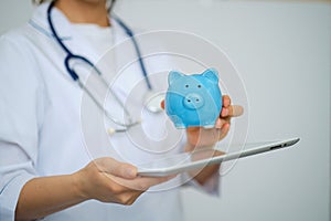 Female Doctor with Stethoscope Holding Piggy Bank and tablet computer. Perfect medical service in clinic. Modern