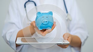 Female Doctor with Stethoscope Holding Piggy Bank and tablet computer. Perfect medical service in clinic. Modern