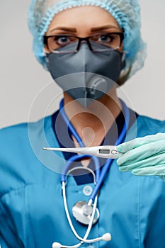 Female doctor with stethoscope wearing protective mask and latex gloves over light grey background holding thermometer