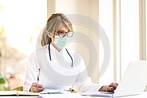 Female doctor wearing face mask while working on laptop in doctor`s room photo