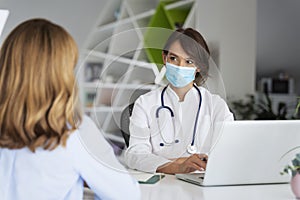 Female doctor wearing face mask and consulting her patient while sitting at desk in doctor`s office photo