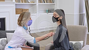 Female doctor wearing face protective mask checking patient lungs with stethoscope while visiting client at home