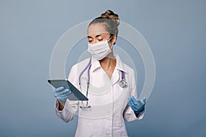 Female doctor wearing protective mask and latex gloves holding stethoscope and tablet on blue background