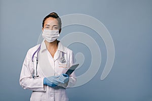 Female doctor wearing protective mask and latex gloves holding stethoscope and tablet on blue background