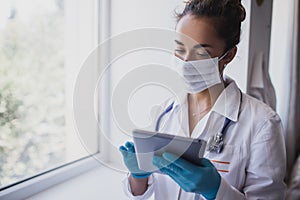 Female doctor wearing protective mask and latex gloves holding stethoscope and tablet, standing near window in a hospital