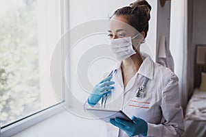 Female doctor wearing protective mask and latex gloves holding stethoscope and tablet, standing near window in a hospital