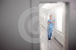 Female Doctor Wearing Scrubs In Hospital Corridor Using Digital Tablet Viewed Through Window In Door