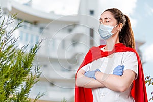 A female doctor wearing a surgical mask and rubber gloves poses with her arms crossed and a super hero's cape on her back