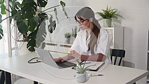 Female doctor in white coat and glasses typing on laptop computer at hospital. Woman medical worker working at the clinic office.