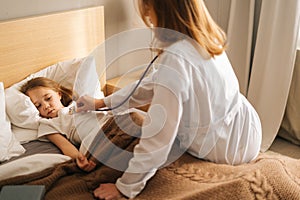 Female doctor in white coat listening to heartbeat with stethoscope to check up little sick girl lying in bed at