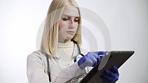 Female doctor in white coat, protective glasses and stethoscope over her neck using tablet at desk while working in