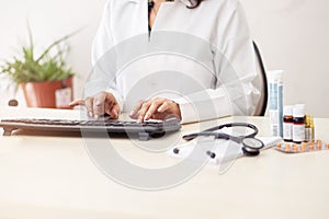 Female doctor in white lab coat,hands typing on laptop computer keyboard with medical stethoscope and  medicine on the mirror desk