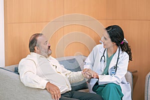 Female doctor in white medical coat and patient discussing something, holding his hand and smiling while sitting on sofa.
