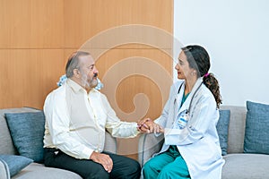 Female doctor in white medical coat and patient discussing something, holding his hand and smiling while sitting on sofa. Medicine