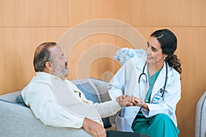 Female doctor in white medical coat and patient discussing something, holding his hand and smiling while sitting on sofa. Medicine