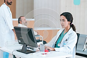 female doctor in white medical coat working with laptop computer, sitting at the table near the window in hospital office, looking